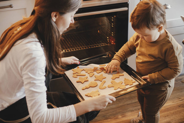 Carino piccola figlia e madre cuocere biscotti di pan di zenzero di Natale nella moderna cucina scandinava. Bella bambina e mamma che tengono il vassoio con i biscotti. Preparativi per le vacanze in famiglia, cucina natalizia - Foto, immagini