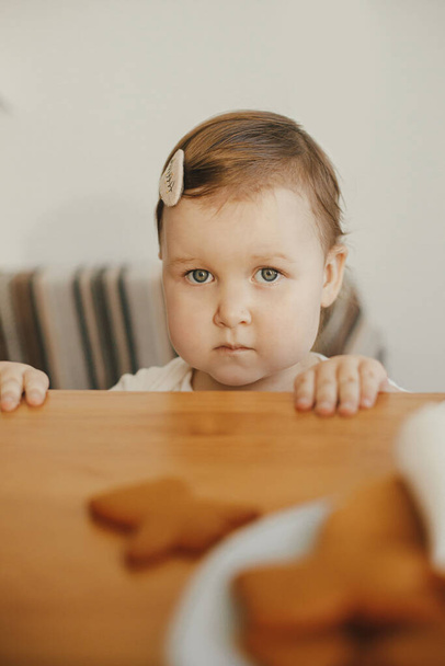 Aranyos kislány nézi a frissen sült mézeskalács sütit a fa asztalon közelről. Hiteles pillanat, ünnepi előkészületek. Imádnivaló kisbaba lány csinál karácsonyi sütit anyával - Fotó, kép
