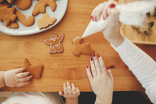 Cute little daughter and mother decorating christmas gingerbread cookies with icing on wooden table, authentic lovely moment. Family time together, holiday preparations. Top view - Photo, Image