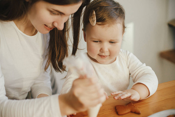 Sevimli küçük kız ve anne birlikte Noel kurabiyelerini modern odadaki ahşap masada kremayla süslüyorlar. Anne kızı otantik güzel anlar. Tatil hazırlıkları - Fotoğraf, Görsel