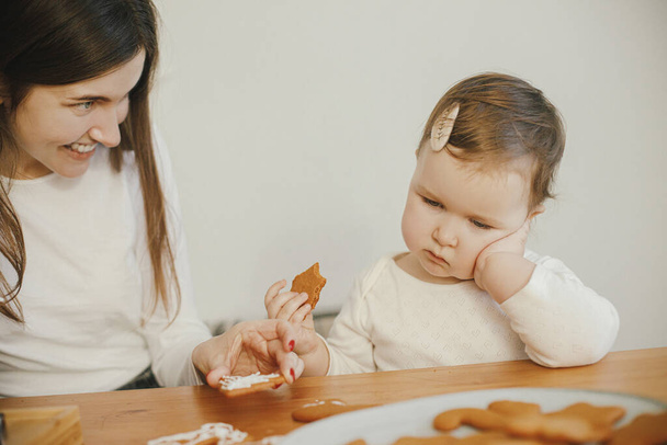Leuke kleine dochter en moeder samen decoreren kerst peperkoek koekjes met glazuur op houten tafel in de moderne kamer. Mama dochter authentieke mooie momenten. Voorbereidingen - Foto, afbeelding
