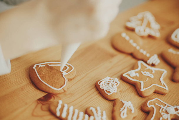 Decorating christmas gingerbread cookies with icing on wooden table close up. Hands decorating baked christmas cookies with sugar frosting. Xmas holiday preparations, atmospheric time - Photo, Image