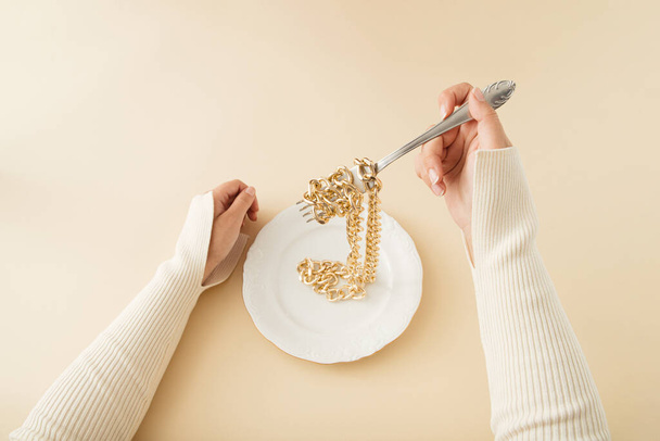 Kobieta zjada złoty łańcuch w talerzu jak spaghetti. Pastelowe, beżowe tło.Artystyczna koncepcja retro. - Zdjęcie, obraz