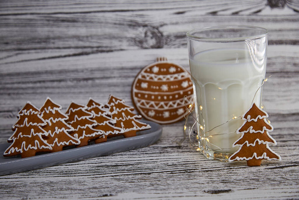 かわいいクリスマスの伝統。木製のテーブルの上に手作りの新鮮なクッキーとミルクの暖かいガラス。テーブルセッティング。お祭りのクリスマスムードとガーランドと審美的な雰囲気。スペースのコピー. - 写真・画像