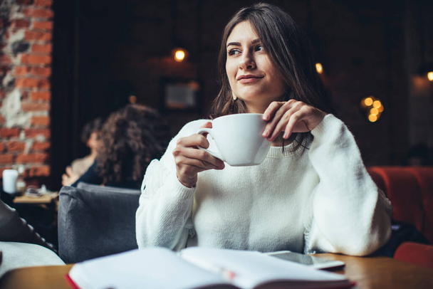 Positive female remote worker looking away while sitting at wooden table in cafeteria with notebook and having coffee break during workday - Foto, Imagem