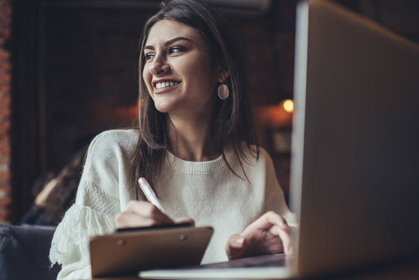 Young female remote worker with toothy smile looking away while sitting in cafeteria taking notes on clipboard and browsing laptop - Foto, Imagem