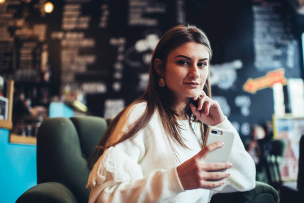 Wistful female in casual clothes with hand at chin looking at camera while sitting in armchair and surfing internet on mobile phone - Zdjęcie, obraz
