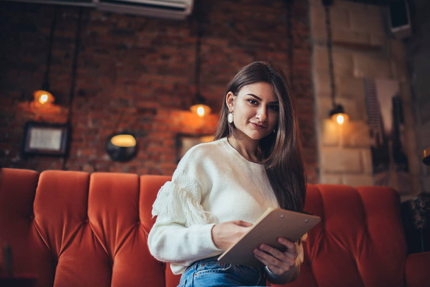 Young female remote worker in casual outfit with long hair looking at camera while sitting on sofa with clipboard while working in cafe - Фото, изображение