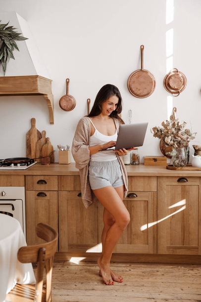 Happy young woman freelancer stands in the kitchen and holds a laptop in her hands - Φωτογραφία, εικόνα