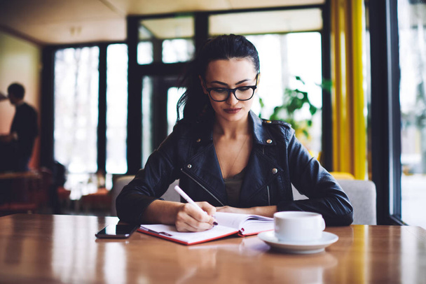 Young focused female student in black casual outfit with eyeglasses looking down while sitting at table with notebook and writing notes in cafeteria - Photo, Image