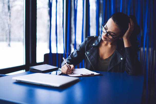 Thoughtful female entrepreneur with black hair in  eyeglasses sitting at blue table and leaning on hand while writing information on paper in clipboard in light room in daytime - Foto, Imagem