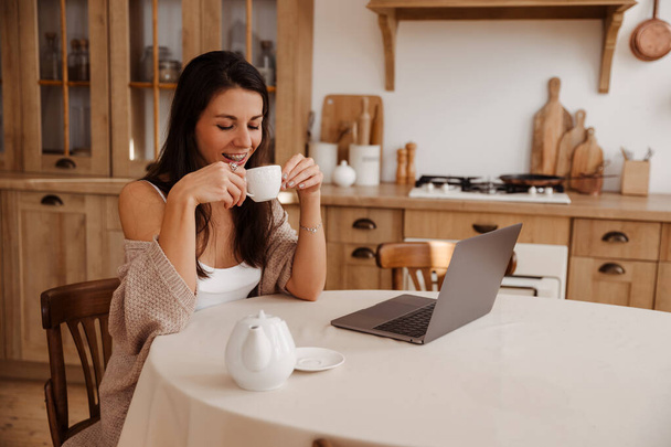 Onnellinen nuori nainen juo kahvia ja työskentelee läppärissä keittiössä - Valokuva, kuva
