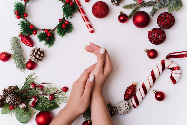 Női kéz fehér tej manikűr fehér alapon karácsonyfa ágak piros dekoráció. Felülről nézve, lapos fekvésű - Fotó, kép