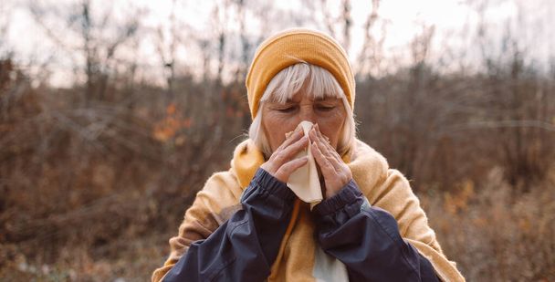 Άρρωστη ενήλικη γυναίκα βήχει και φυσάει τη μύτη της στο πάρκο κατά τη διάρκεια της φθινοπωρινής περιόδου σε μια κρύα ημέρα του φθινοπώρου. - Φωτογραφία, εικόνα