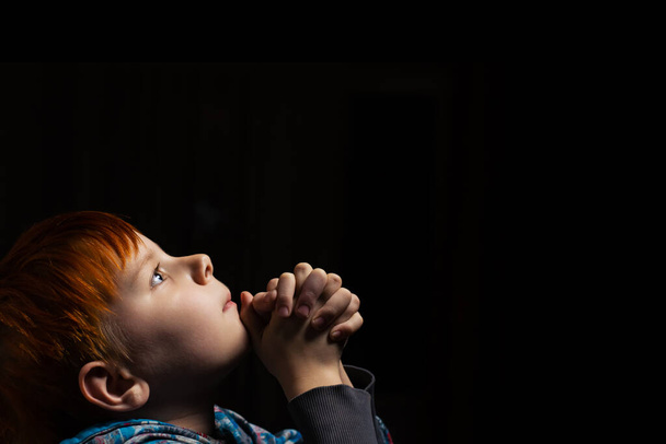 Prayer of a child in the dark. The red-haired boy is praying. Side view. - Foto, imagen
