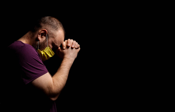 A man in a medical mask is praying on a black background. Turning to God for help from coronavirus concept. - Фото, изображение