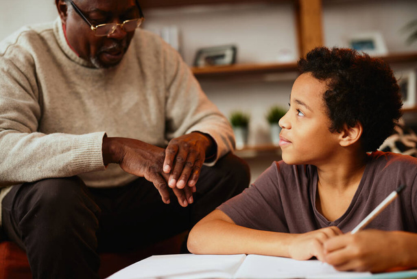 Un abuelo afroamericano está sentado en casa con su nieto y le ayuda con los deberes.. - Foto, Imagen