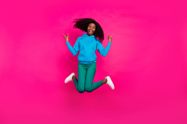 Photo of funky pretty dark skin lady wear blue hoodie jumping high showing hard rock sign smiling isolated pink color background - Foto, Imagem
