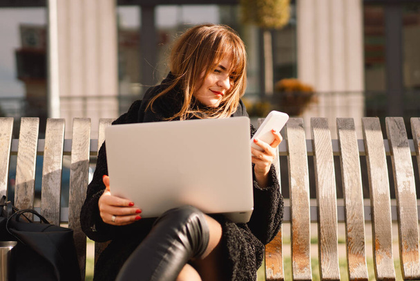 Een vrouw zit op een bankje in de stad, gebruikt een computer en een telefoon. Freelance vrouw. - Foto, afbeelding