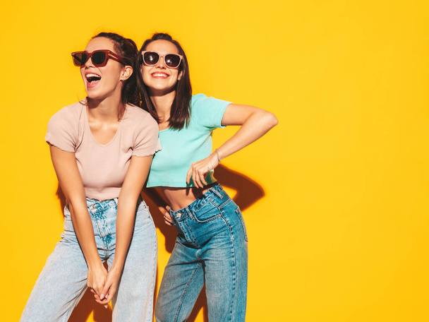 Two young beautiful smiling blond hipster female in trendy summer clothes. Sexy carefree women posing near yellow wall in studio. Positive models having fun. Cheerful and happy. In sunglasses - Zdjęcie, obraz