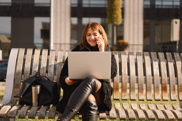 Женщина сидит на скамейке в городе, пользуется компьютером и телефоном. Фрилансер. - Фото, изображение