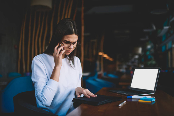 Young concentrated woman remote worker with eyeglasses thoughtfully looking down while sitting at table with laptop and notepad having conversation on mobile phone - Foto, afbeelding