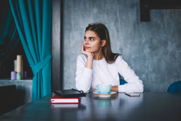 Young female in white jumper leaning on hand sitting at table with cup of hot drink and thinking looking away in cafe - Foto, Bild