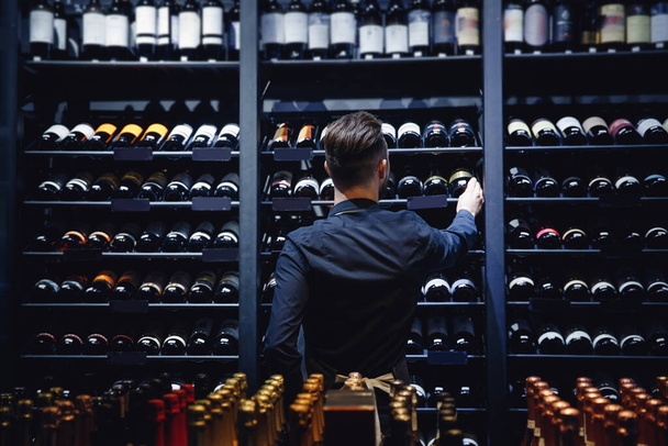 Sommelier Barista all'enoteca pieno di bottiglie con bevande alcoliche, vista posteriore - Foto, immagini
