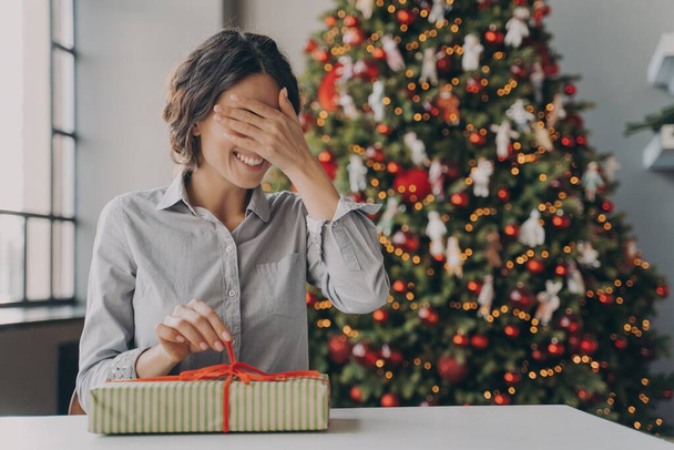 Aufgeregte Europäerin bekommt Weihnachtsüberraschung, sitzt am Schreibtisch neben geschmücktem Weihnachtsbaum zu Hause - Foto, Bild
