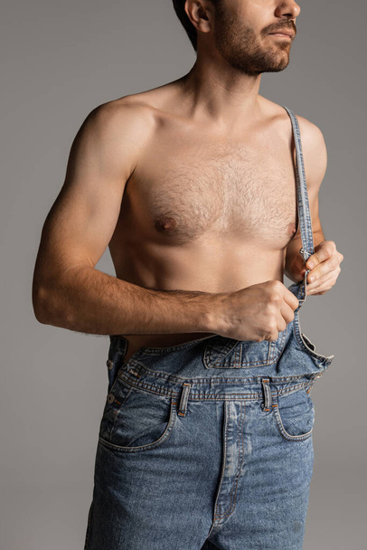 Cropped portrait of young handsome bearded shirtless man in jeans posing isolated on gray studio background. - Foto, Bild