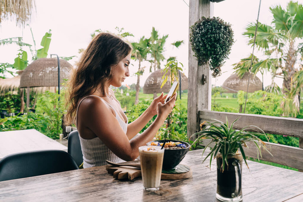 Side view of focused ethnic female browsing cellphone while sitting at table with tasty food and coffee during breakfast time in outdoor cafe - Foto, imagen