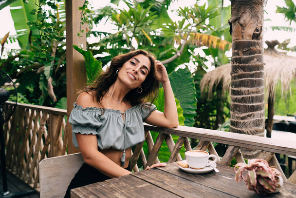 Cheerful young ethnic woman in trendy summer wear leaning on balcony railing with elbow and touching hair while having coffe break in outdoor cafe in tropical park - Foto, imagen