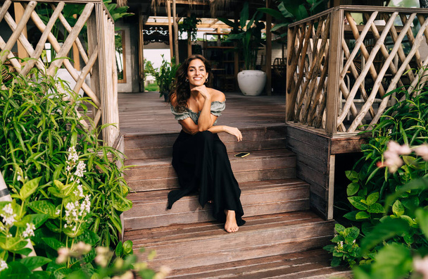 Positive barefoot female in stylish outfit sitting alone on steps leaning head on hand while resting among blooming plants near fences of wooden terrace of comfortable cafe in sunny day - Foto, Bild