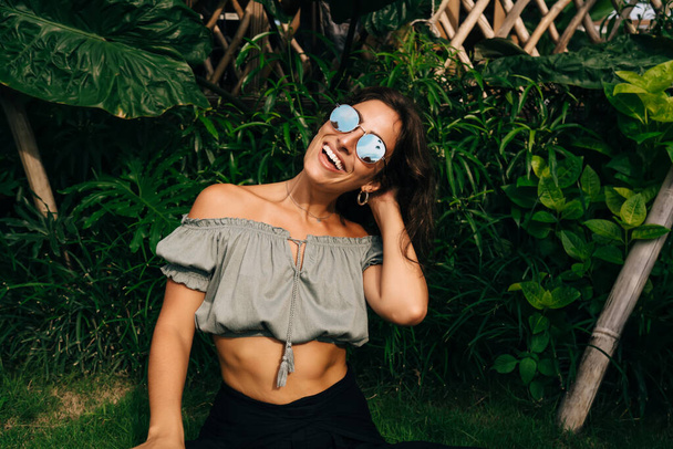 Happy young fit ethnic female in trendy outfit and sunglasses touching hair and smiling while spending free time in green garden during summer holidays - Photo, image