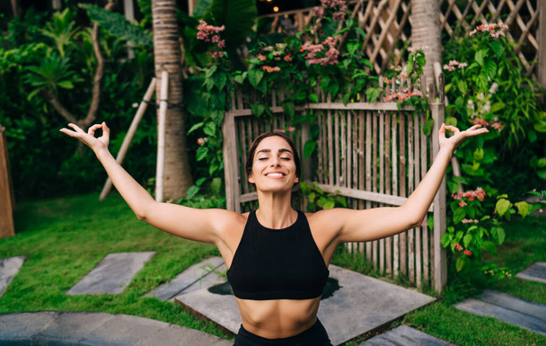 Happy female yogi practicing hatha during leisure time for mental harmony keeping healthy lifestyle, cheerful Caucasian woman in black top feeling freedom and pleasure while recreating outdoors - Фото, изображение