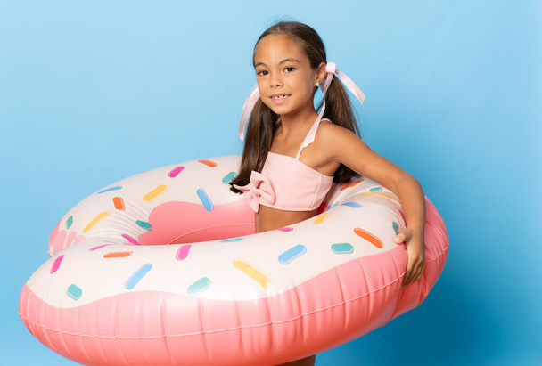 Cheerful little girl wearing swimsuit standing isolated over blue background, playing with inflatable ring - Photo, Image