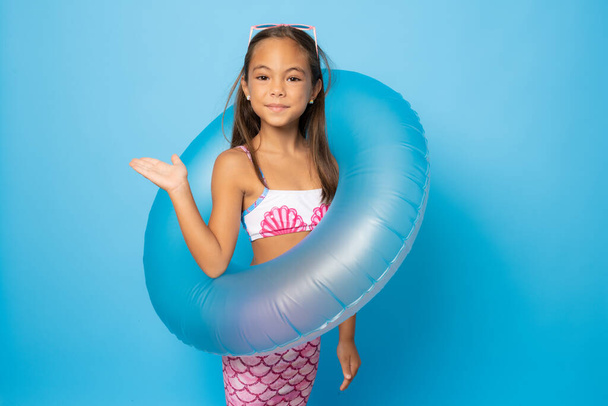 Cheerful little girl wearing swimsuit standing isolated over blue background, playing with inflatable ring - Photo, Image