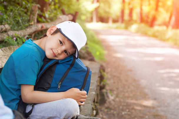 Мальчик в рюкзаке и кепке сидит у дороги и ждет автобус.. - Фото, изображение