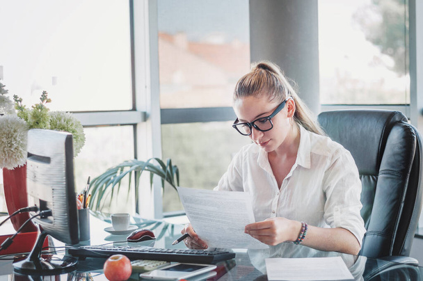 Portret van een jonge zakenvrouw in een bril die op haar werkplek zit en documenten leest. Kantoormedewerker op moderne kantoorwerkplek. Bedrijfsconcept.  - Foto, afbeelding