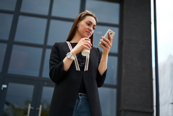Caucasian female blogger with takeaway caffeine beverage connecting to 4g wireless on cellphone device for browsing media content, millennial hipster girl reading web publication on mobile gadget - Foto, imagen