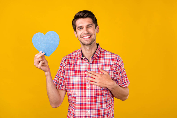Portrait of attractive cheerful guy holding blue card sign touching heart amour isolated over bright yellow color background. - Photo, Image