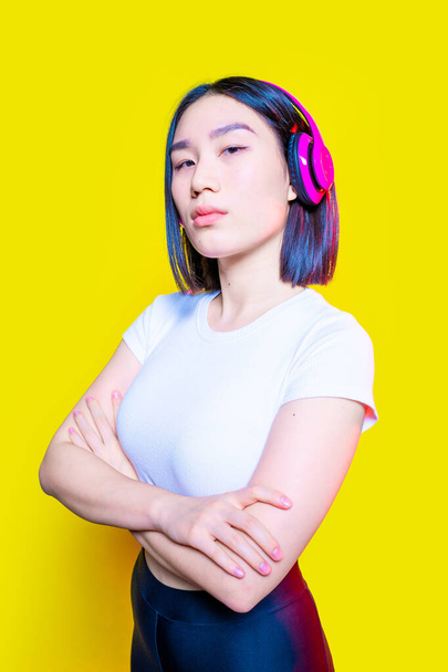 Sportowe młoda azjatycka kobieta pokazuje moc z ramionami złożone na izolowanym żółtym tle patrząc aparat poważne słuchanie muzyki bezprzewodowy słuchawki - Zdjęcie, obraz