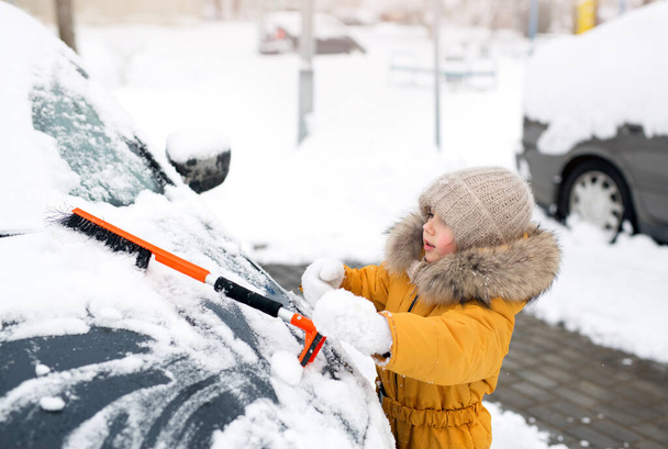 Kind reinigt een auto bedekt met sneeuw en ijs op de parkeerplaats na een sneeuwval. Sneeuwruimen. Schraper voor het verwijderen van ijs uit de auto. Meisje in een oranje winterpak, wanten en een gebreide wollen muts - Foto, afbeelding
