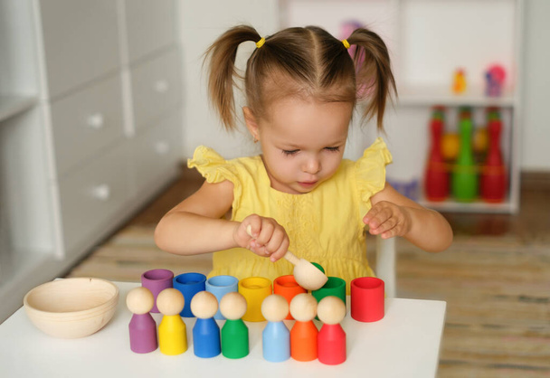 Preschooler kid learns colors and shapes by playing a wooden educational multicolored toy with balls and cups. A focused Child is sitting at a table in the children's room. Two - year - old girl - 写真・画像