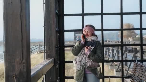 Femme en vêtements chauds sourit et utilise son téléphone sur la plage d'hiver, voyageur à la mer en hors-saison, 4k 60p - Séquence, vidéo