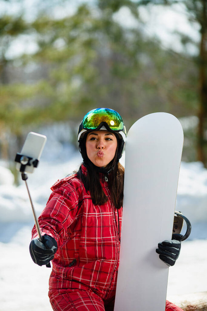 Woman with the snowboard using smartphone on a stick to making selfie - Foto, immagini
