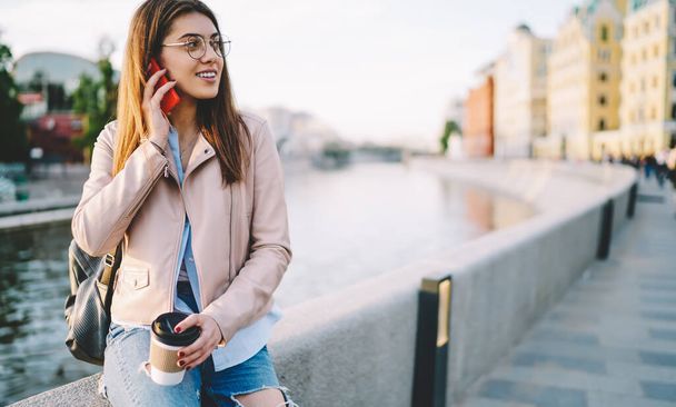 Brunette millennial woman in eyewear and stylish clothing making mobile phone conversation during free time outdoors holding coffee to go, Caucasian tourist calling via cellular using roaming internet - Photo, image
