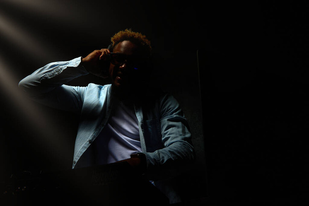 Afrikai-amerikai DJ fejhallgatóval ül egy sötét szobában, kis fénysugarak hullanak rá. DJ egy klubban, színes zenével. - Fotó, kép