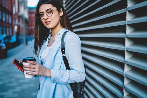 Attractive female tourist with modern mobile device and takeaway coffee cup posing in city, millennial woman using mobile phone and 4G connection for blogging during travel journey on vacations - Photo, image