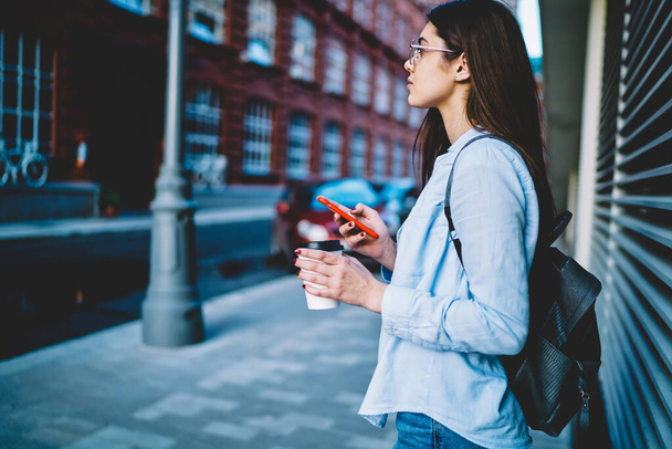 Side view of pensive hipster girl in casual wear thinking about touristic vacations during coffee break,thoughtful female blogger with smartphone device and caffeine beverage feeling pondering in city - Foto, Bild